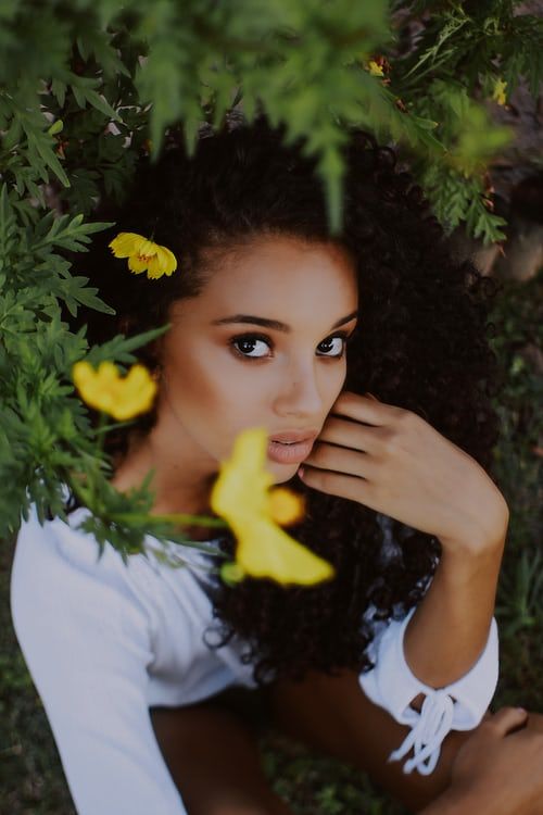Woman with curly hair and yellow flowers in it, looking up. She is in a green, natural setting.