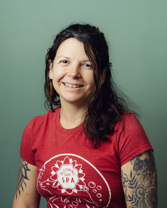 Smiling woman in red T-shirt against a green background, with dark hair and tattooed arms.