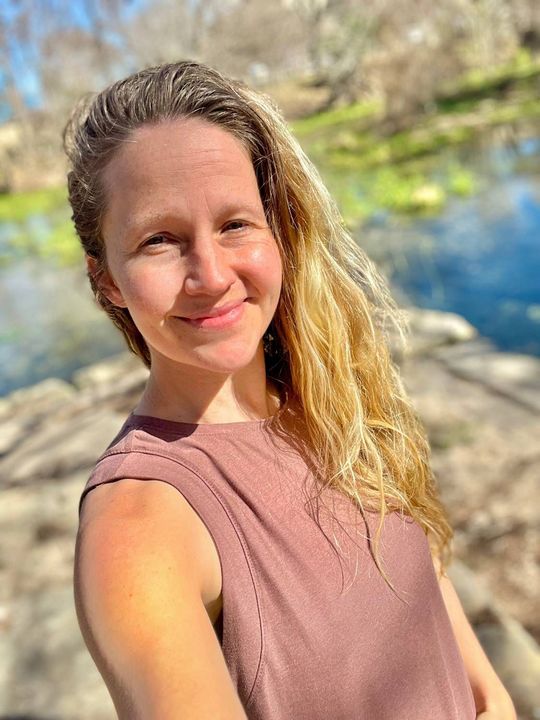 Person in a pink sleeveless top smiling outdoors by a sunlit river.
