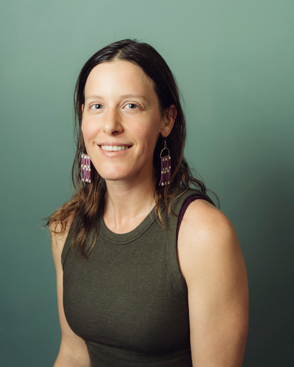 Person with long dark hair and dangling earrings, smiling in a sleeveless green top against a muted green background.