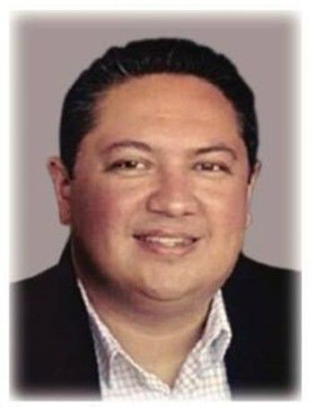 Man in a suit jacket smiles, looking towards the viewer; white shirt visible.