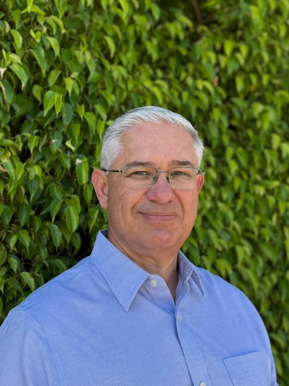 Man with glasses in a blue shirt and dark blazer, smiling at the camera against a blurred background.