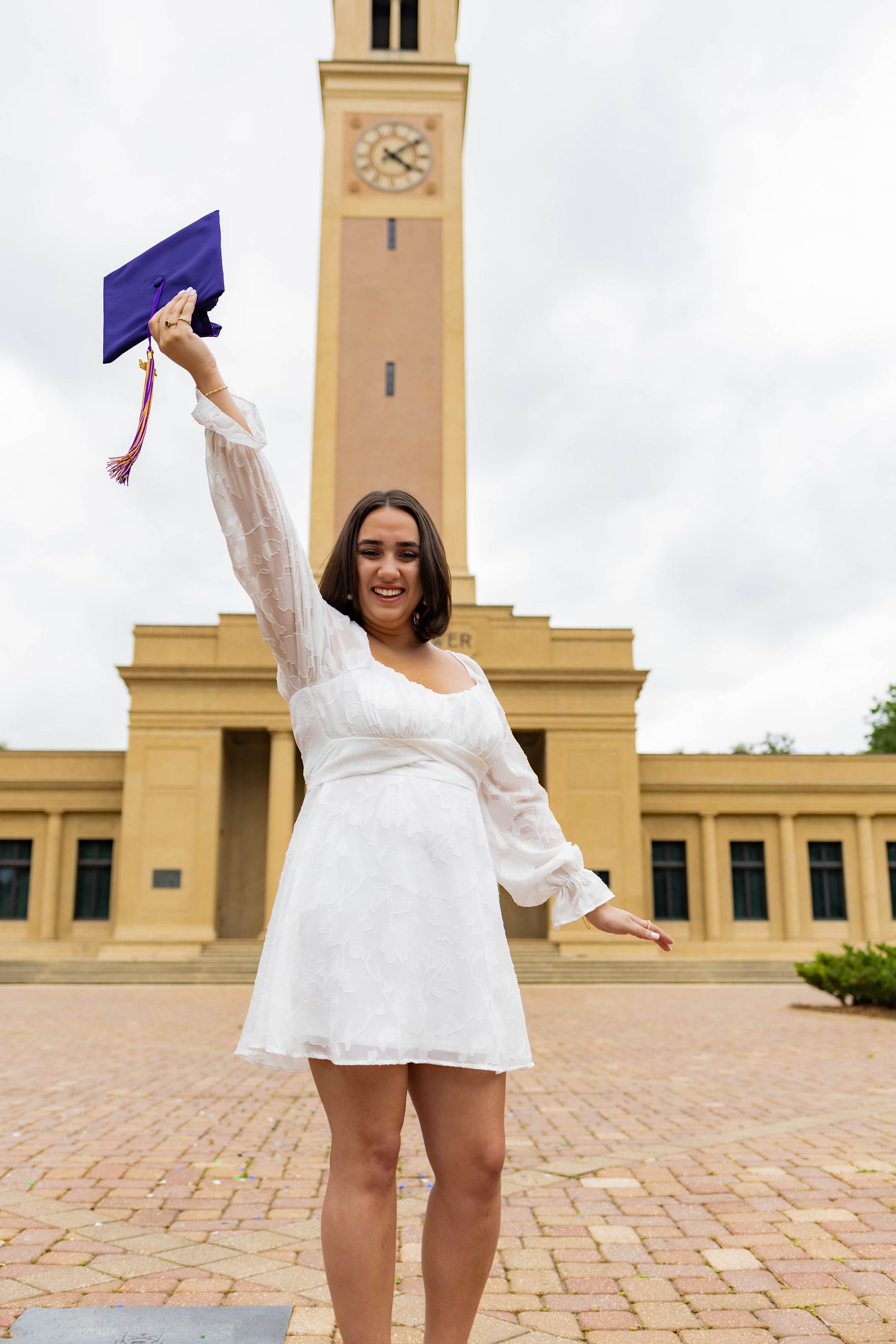 A woman in a white dress is holding a graduation cap in front of a clock tower.