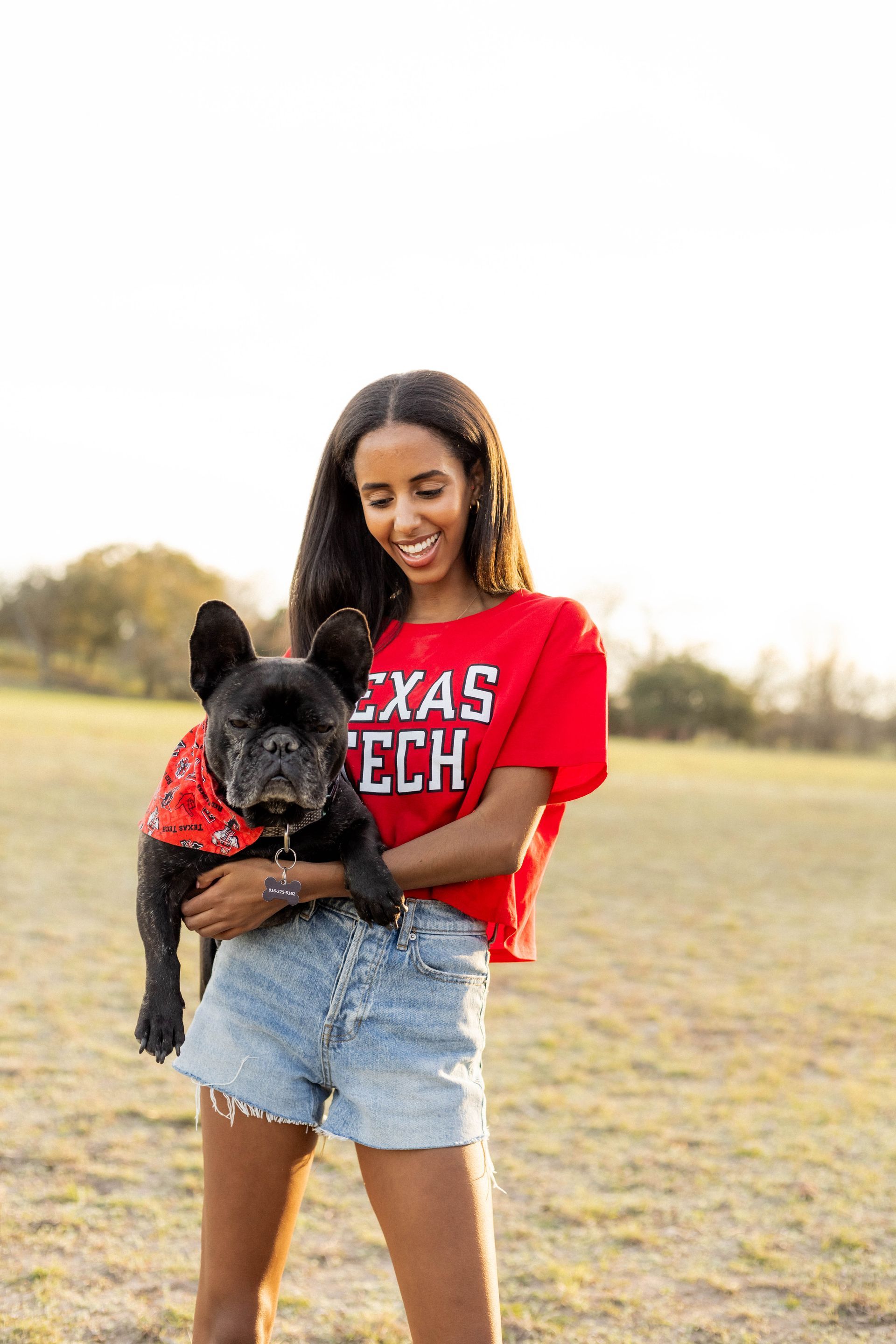 A woman in a red shirt is holding a black dog in a field.