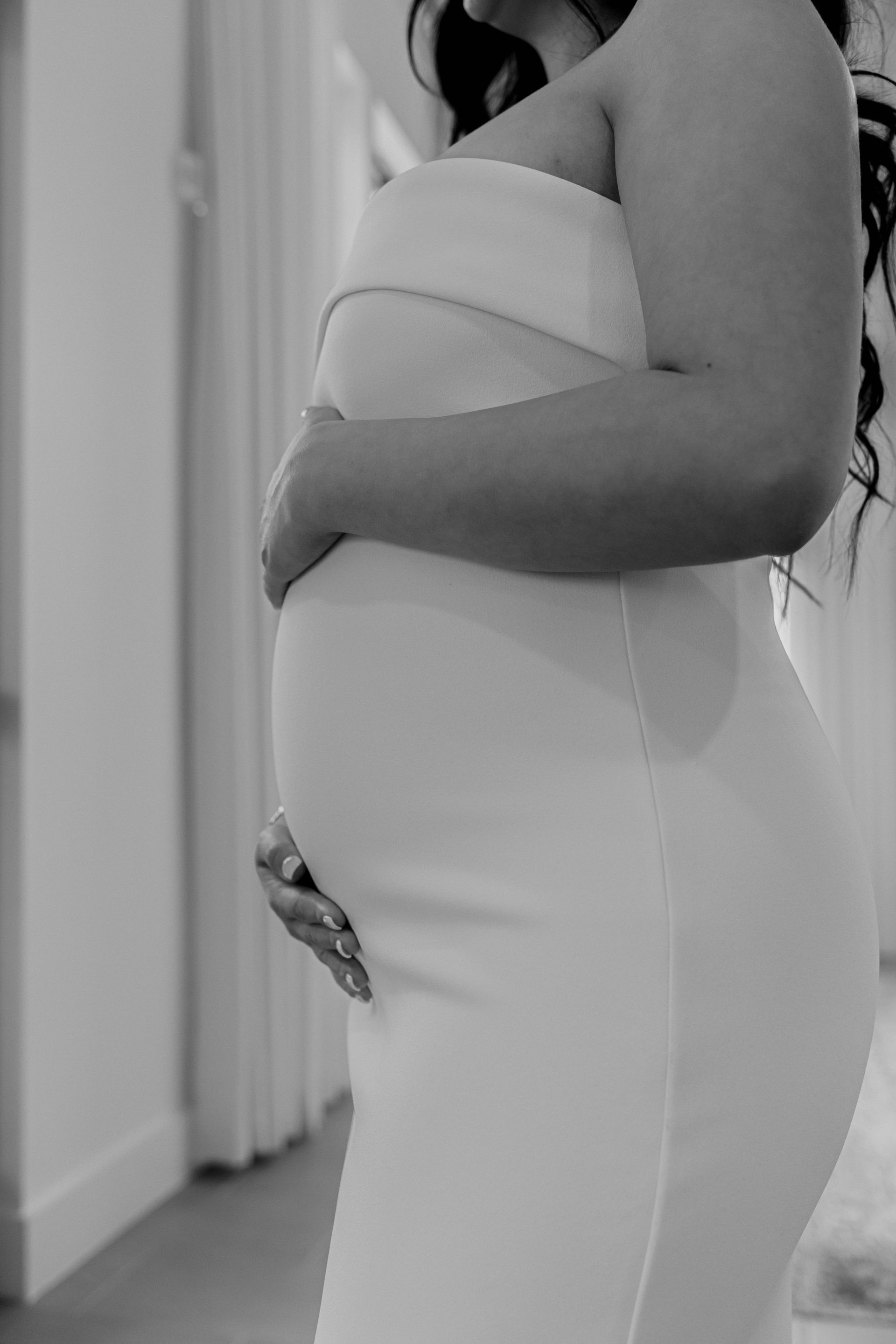 A pregnant woman in a white dress is holding her belly in a black and white photo.