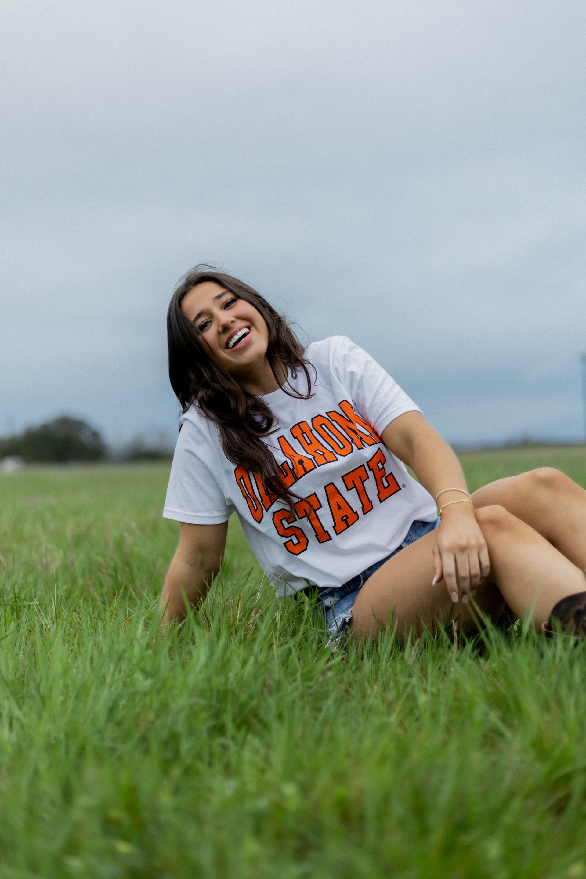 A woman is sitting in the grass wearing a t-shirt that says oklahoma state.