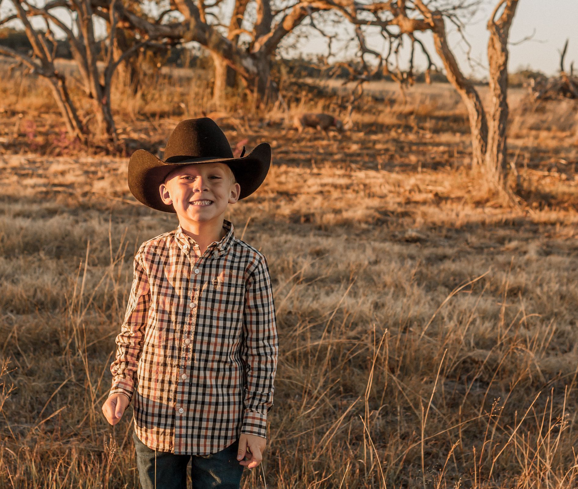 A young boy wearing a cowboy hat and plaid shirt is standing in a field.
