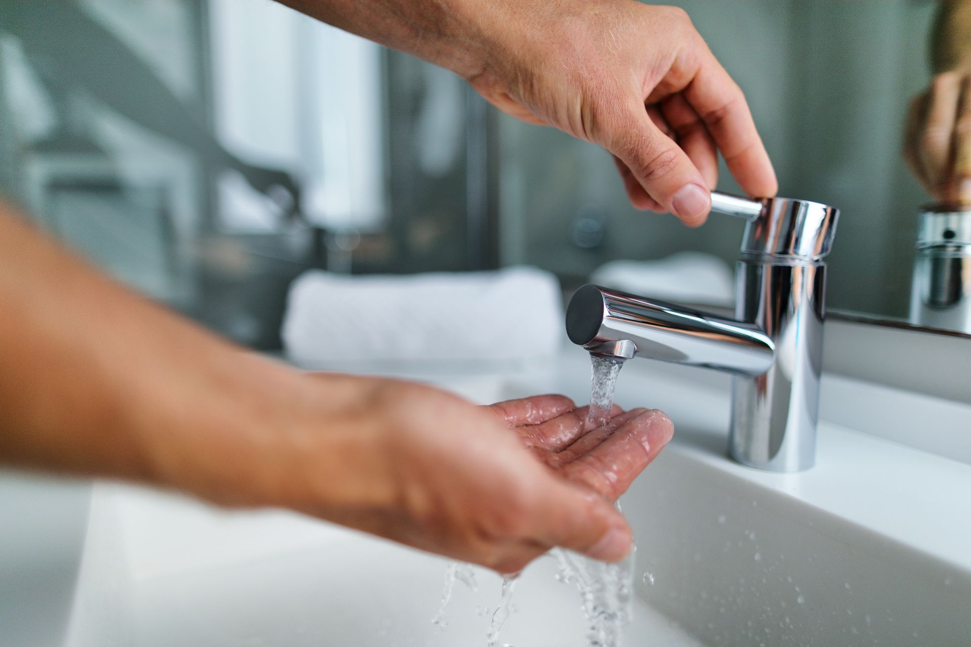 Hand turning a modern faucet while water flows over hands in a bathroom sink.