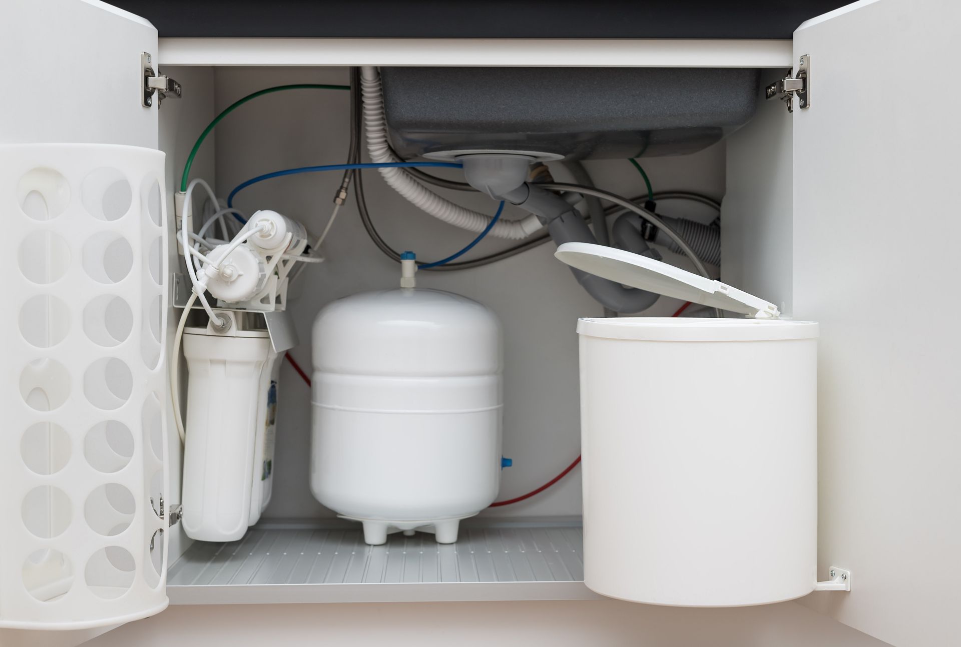 Under-sink water filtration system with storage tank, filters, and connected tubing inside a kitchen cabinet.