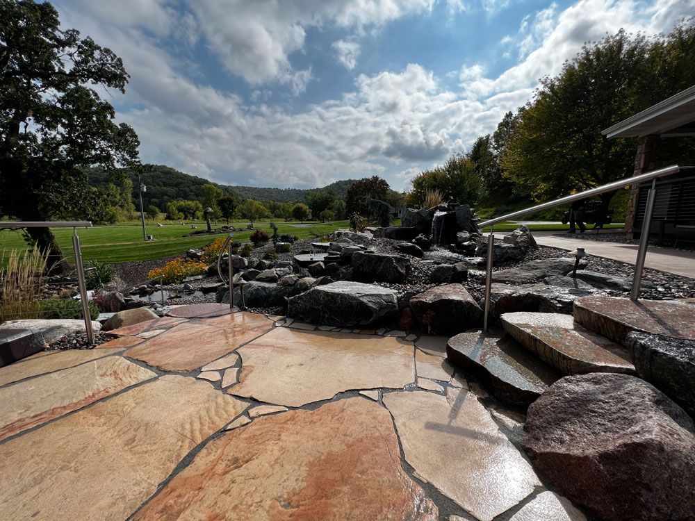 Stone patio with waterfall and view of green valley under a cloudy sky.