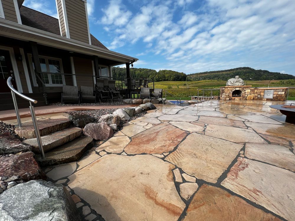 Stone patio with steps and an outdoor kitchen near a house, overlooking a field.