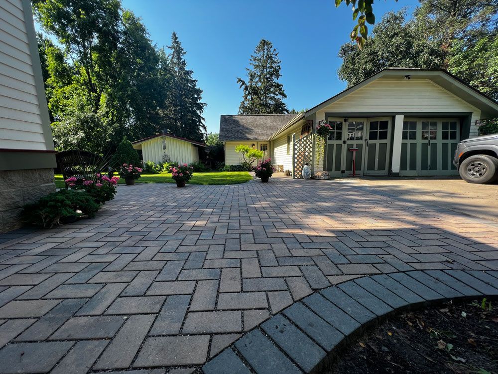 Brick driveway leading to a house with a garage, flower pots, and a shed on a sunny day.