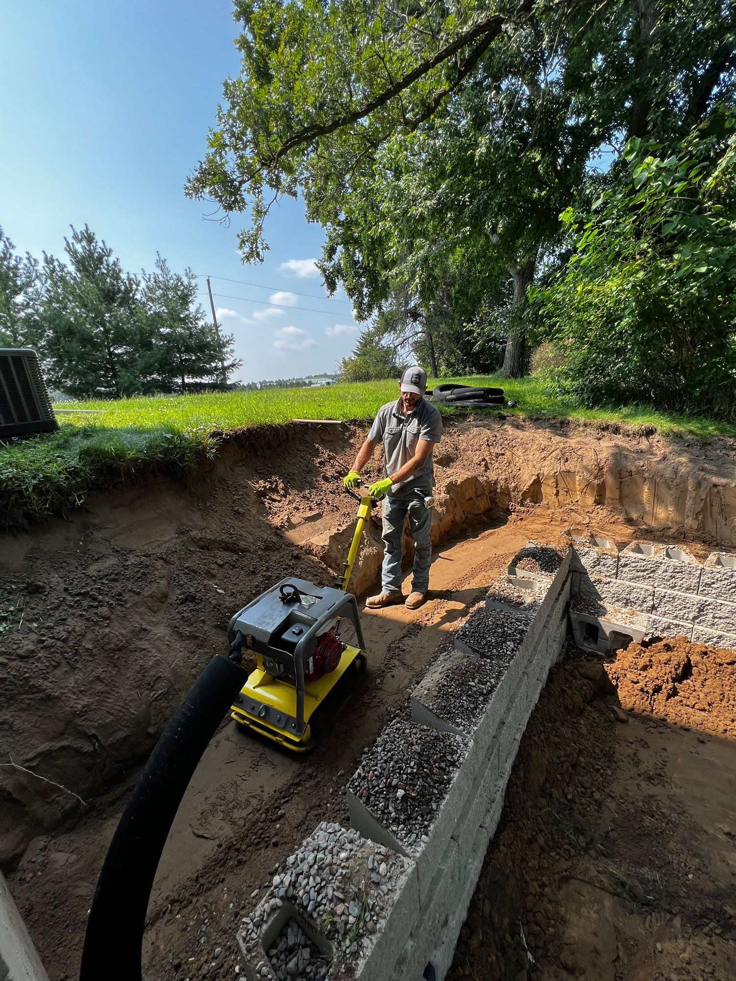 Person operating a soil compactor in a construction site with dirt walls, a retaining wall, and trees.