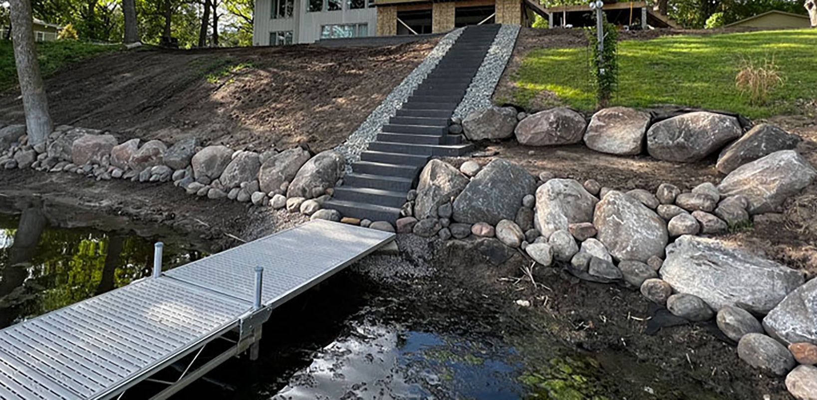 Dock with stairs leading up a sloped bank to a house on a lake. Rocks line the bank.