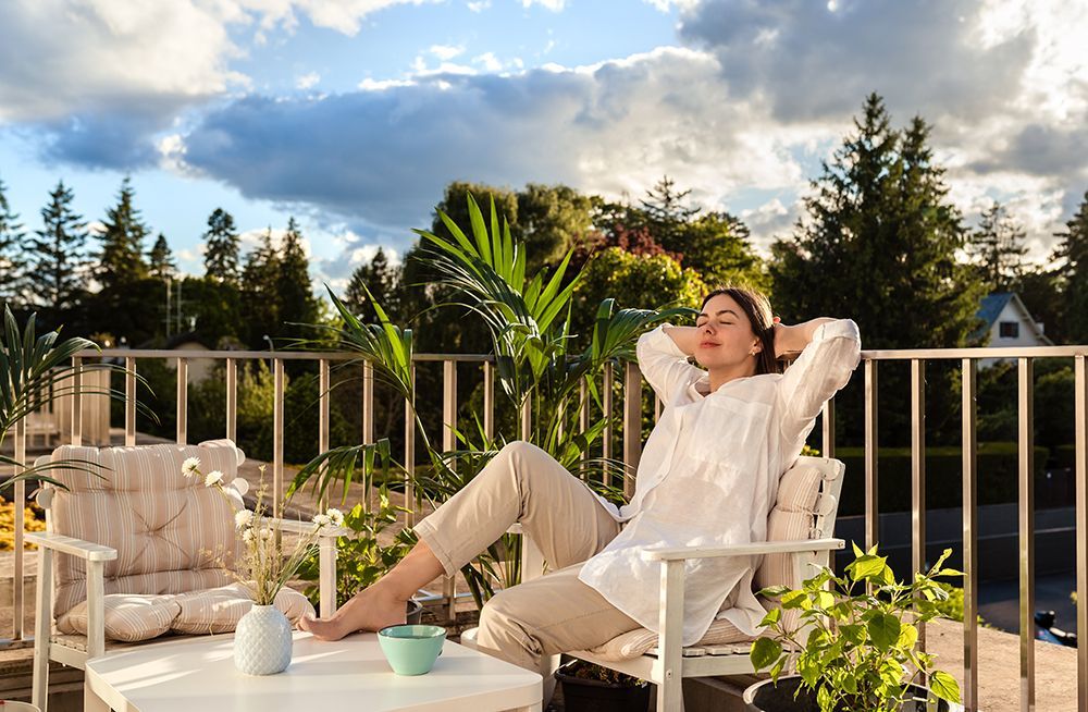 Woman relaxing on a balcony, arms behind head, with trees, sky, and plants visible.
