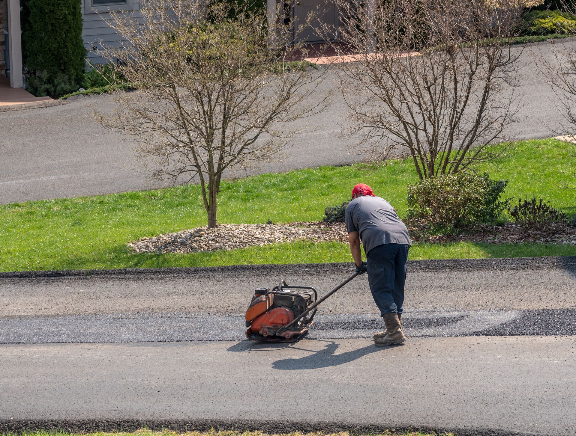 A worker operating a plate vibrator to compact a layer of fresh tarmac, repairing damage on an asphalt street.