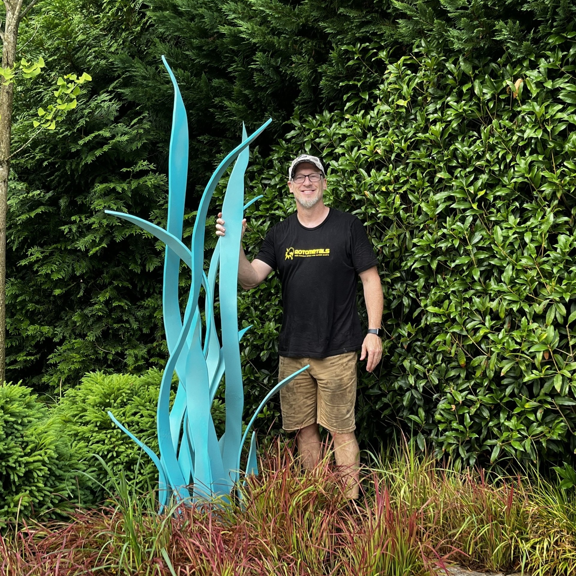 A man is standing next to a large blue sculpture in a garden.