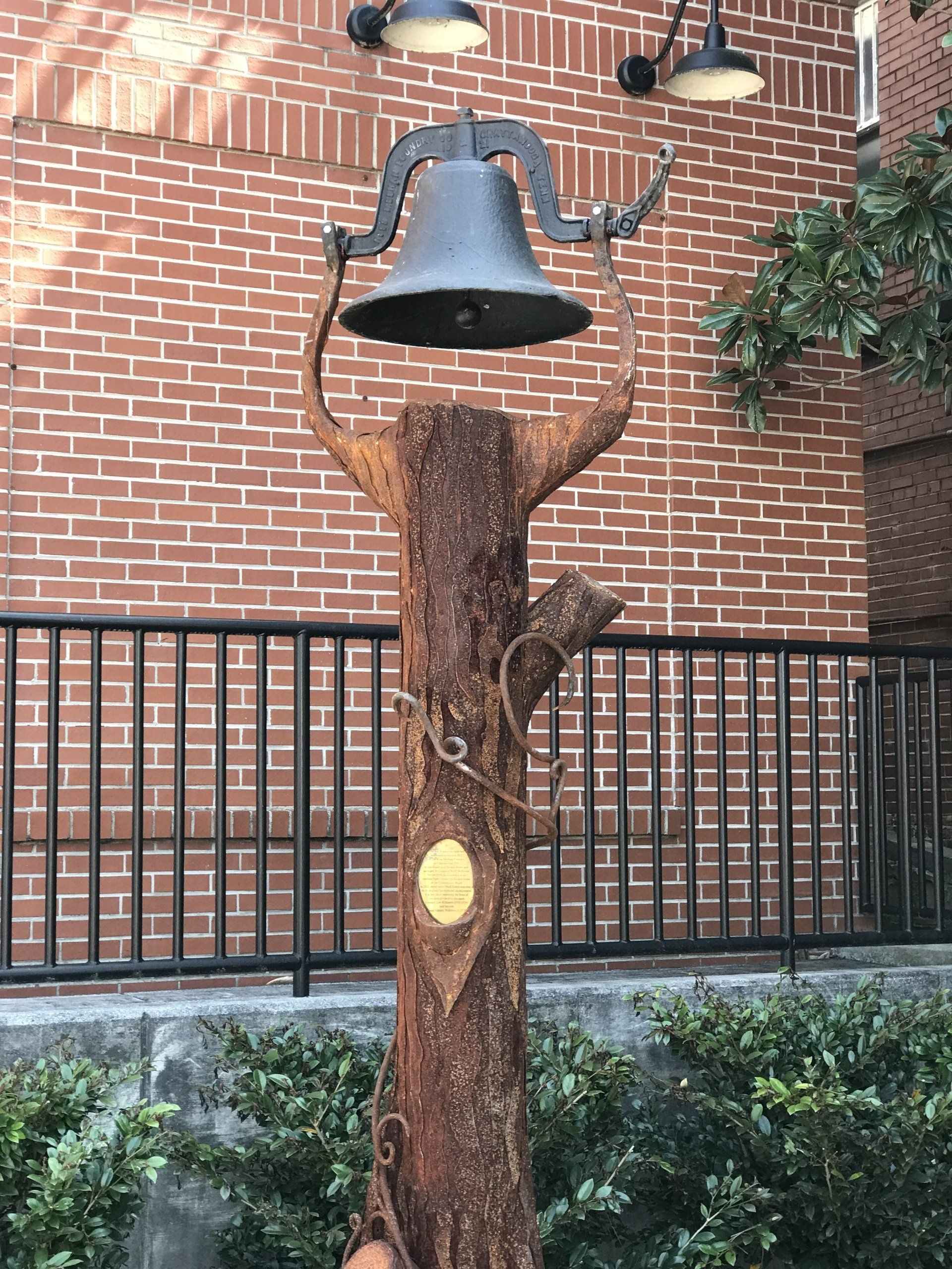 A bell is hanging from a tree stump in front of a brick building.