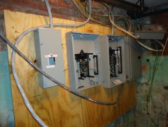 Open electrical breaker panel boxes mounted on a plywood wall with exposed wires.