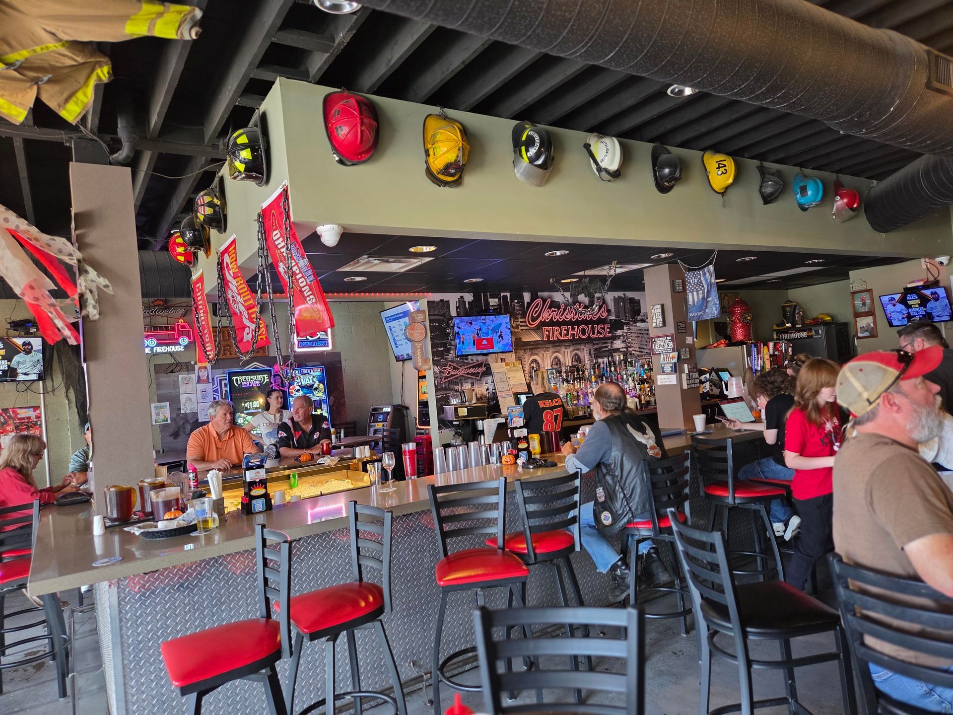 A bar with patrons and staff. Red and black bar stools line the counter. Firefighter helmets are hung above the bar.