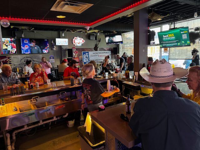 People at a bar, some at counter, others seated. Televisions on walls. Bright interior with red trim.