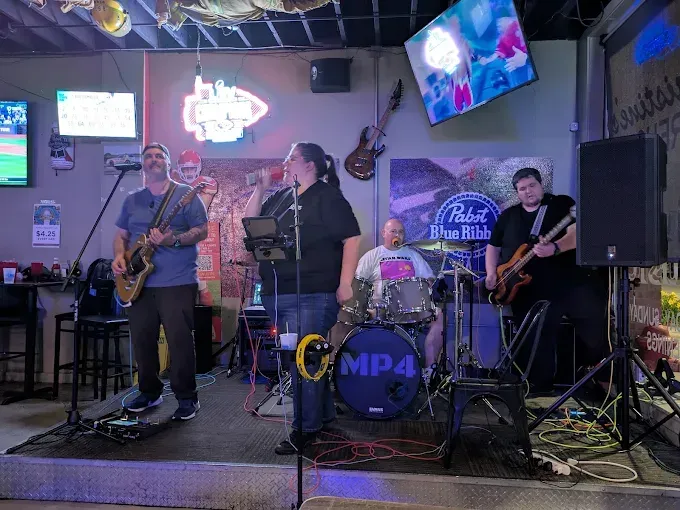 Band performing live at a bar. Musicians play guitar, bass, drums, and sing into a microphone. Dark setting with stage lights.