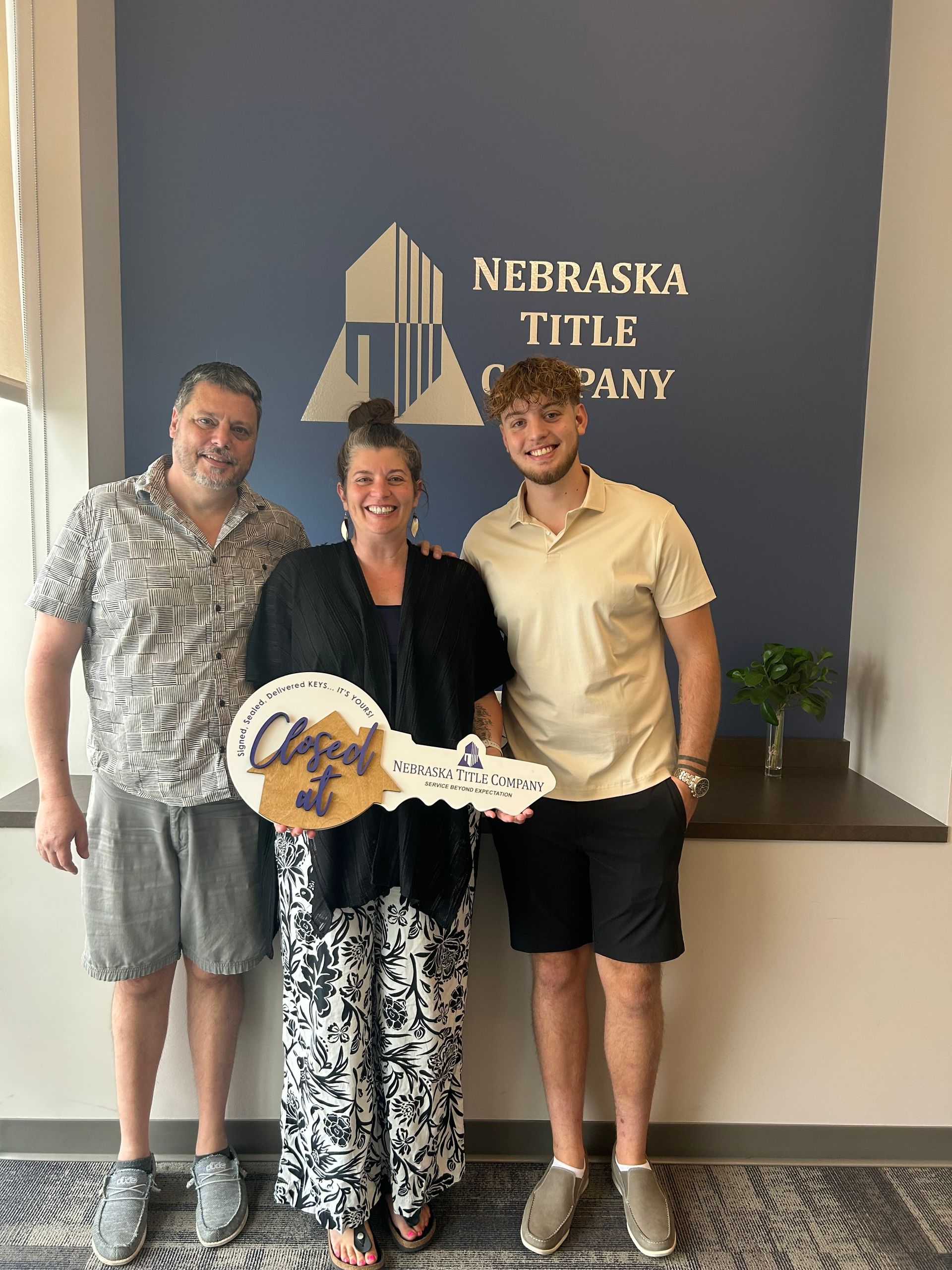 People celebrating a new home purchase. Woman holding a large key, posing with two men in front of a Nebraska Title Co. sign.