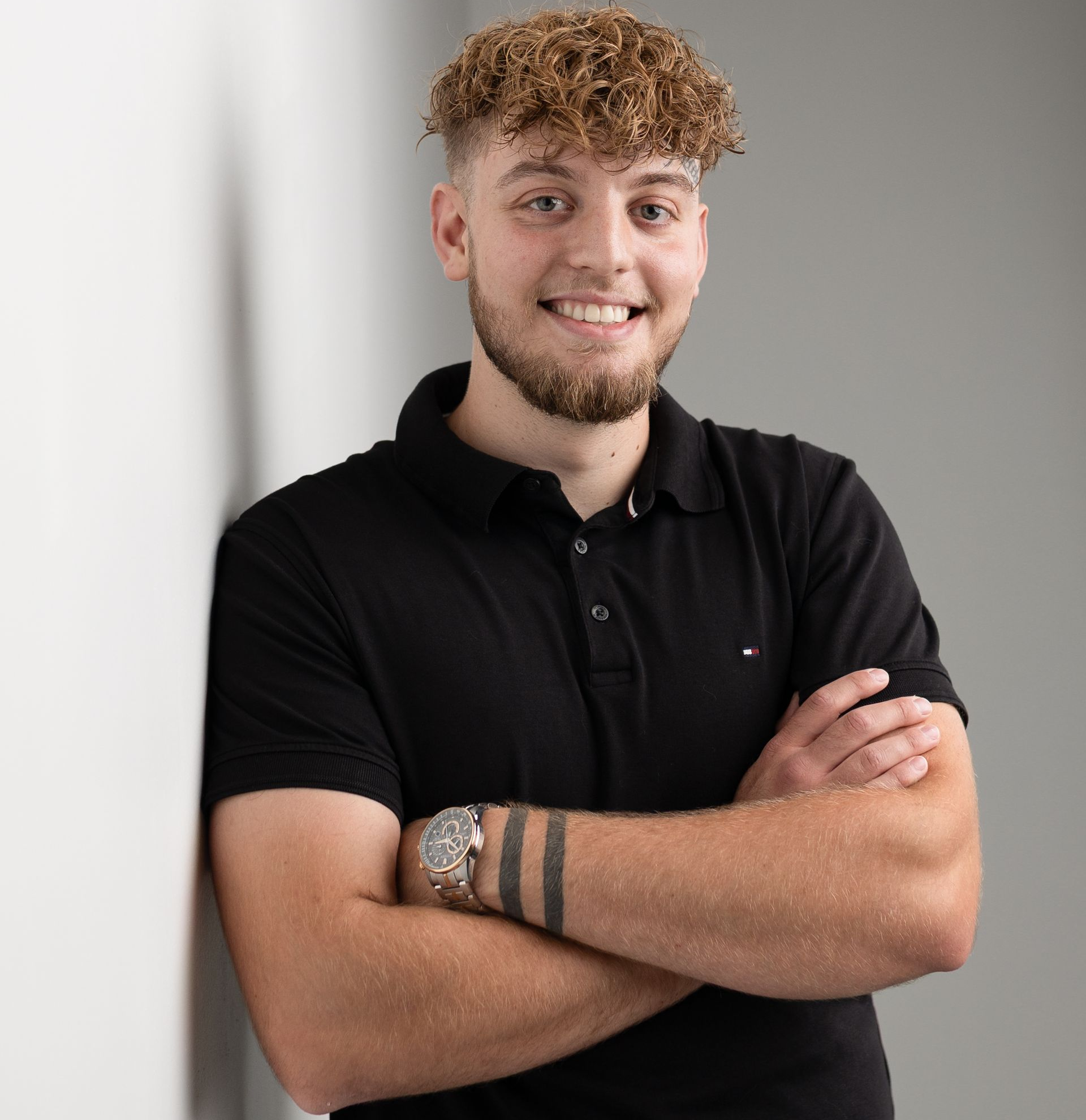 Man with curly hair and beard smiles, arms crossed, wearing a black polo shirt and a watch.