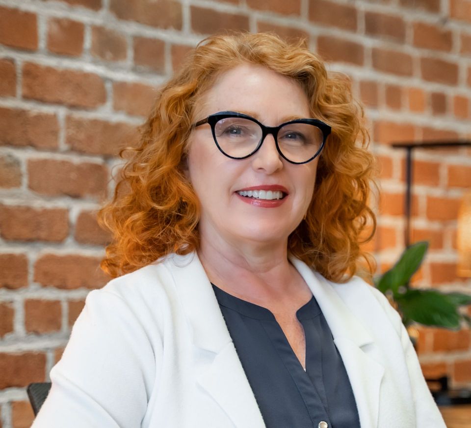 A woman with red hair and glasses is smiling in front of a brick wall.