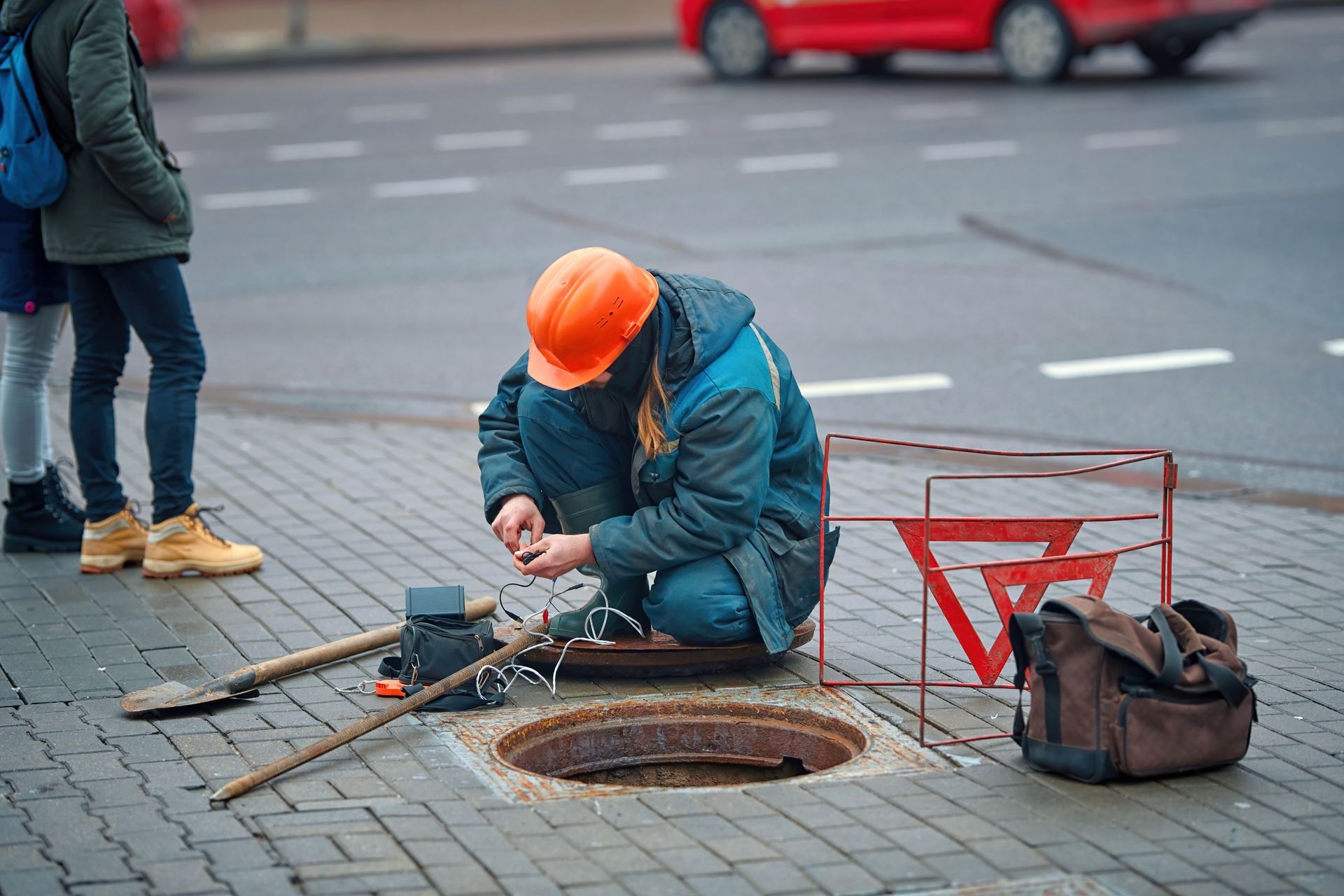 Worker in hard hat crouching by open manhole, handling cables with tools on sidewalk.