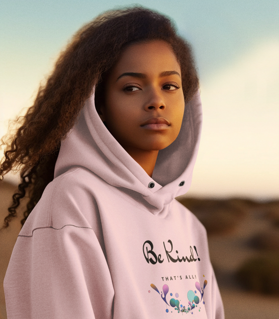 Woman wearing a pink hoodie with “Be Kind” on it, looking thoughtful outdoors.