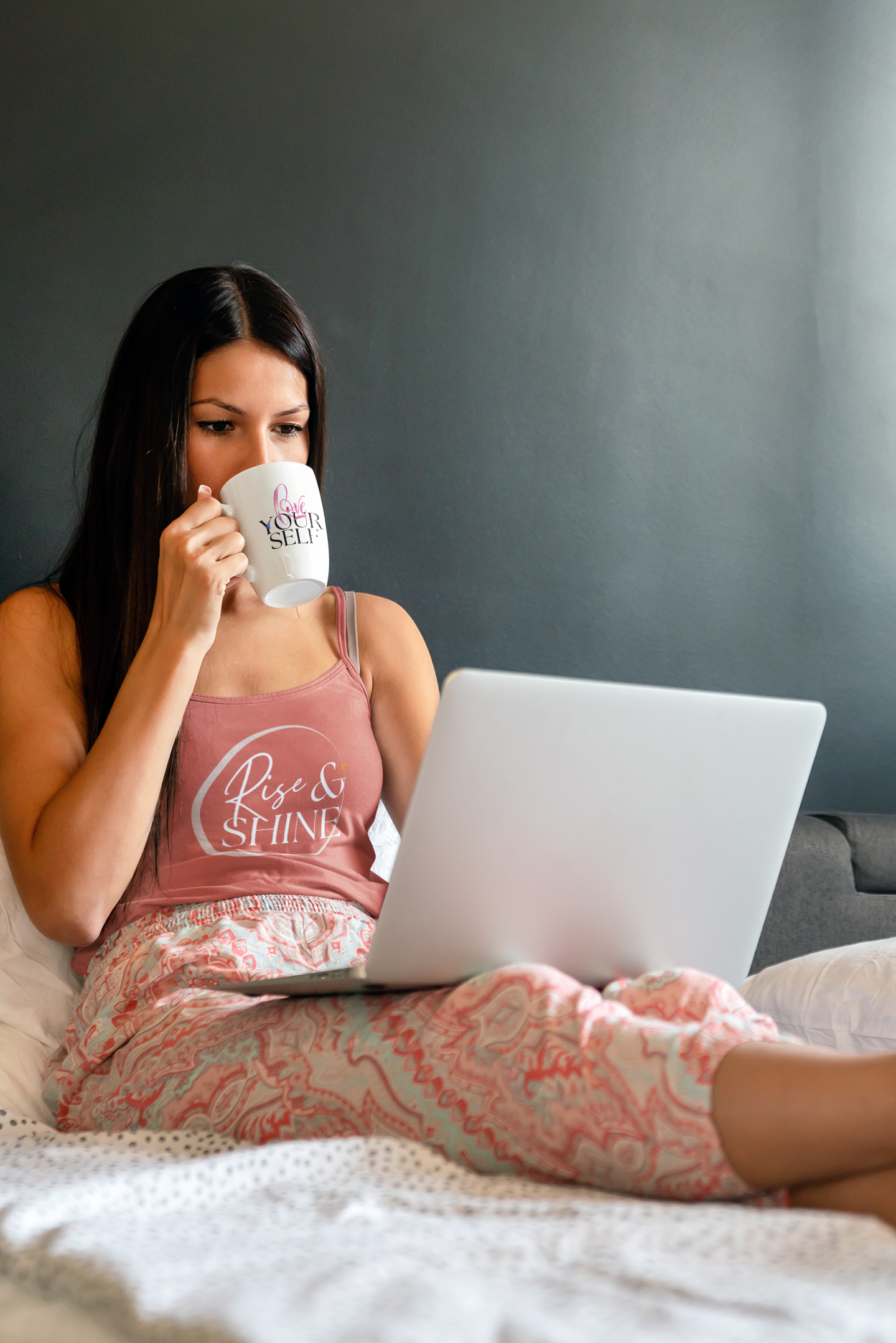 A woman is sitting on a bed drinking coffee and using a laptop computer.