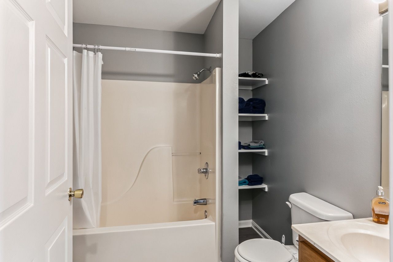 Bathroom with shower, toilet, shelving, and closed door. Gray walls, white fixtures, and a wooden cabinet.