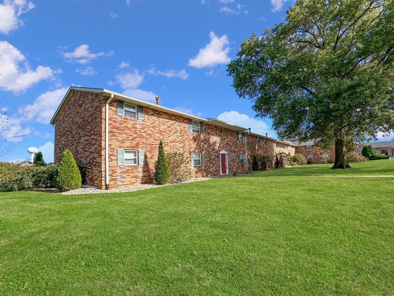 Brick townhouses with green lawn and large tree under a partly cloudy blue sky.