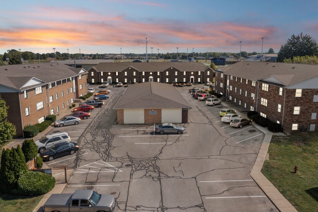 Apartment complex with parking lot, cars, and a central building, set against a sunset sky.