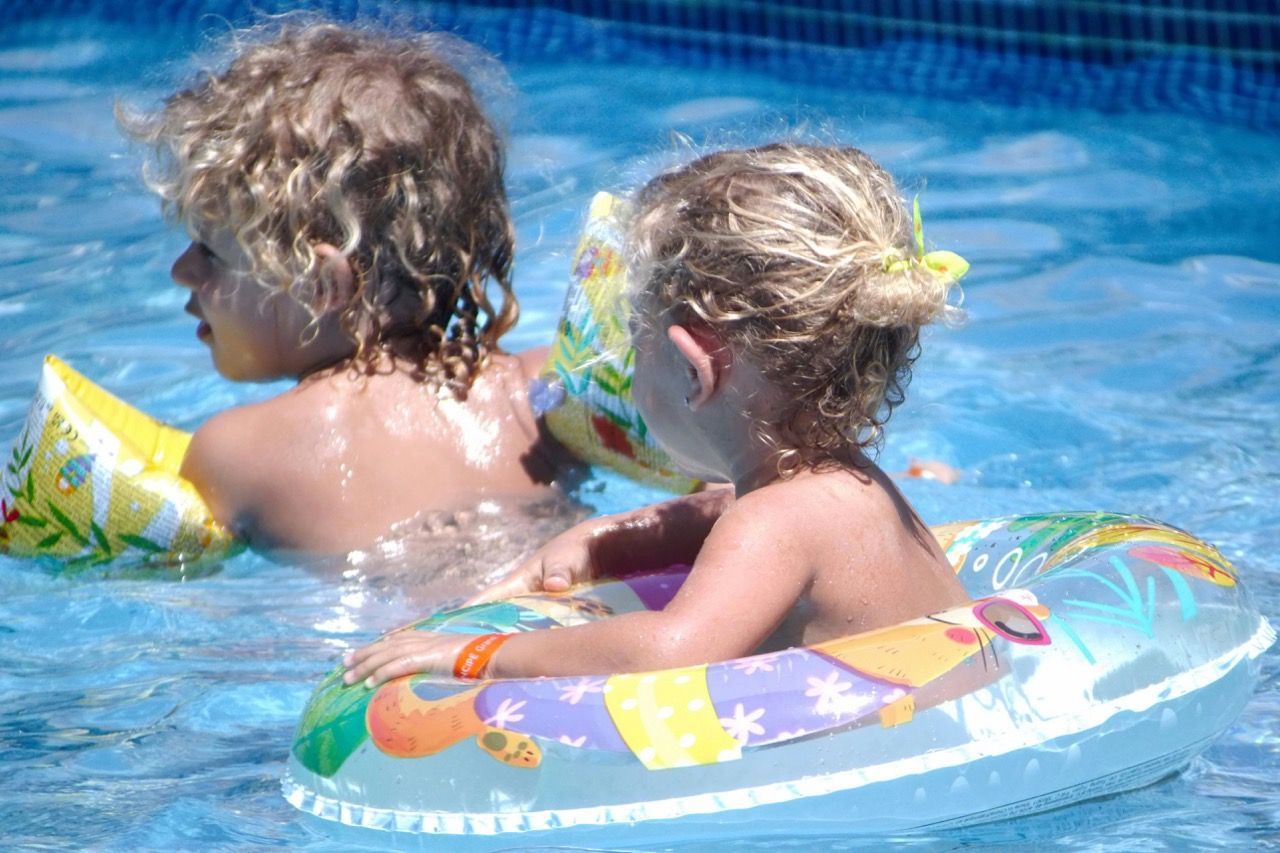 Two children in a pool. One wearing arm floaties, the other in a tube. Both have blond hair, playing in the sun.