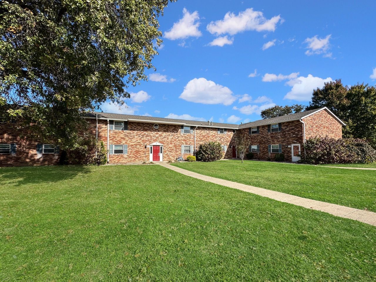 Two-story brick apartment building with a grassy lawn and a blue sky. A red door is visible.