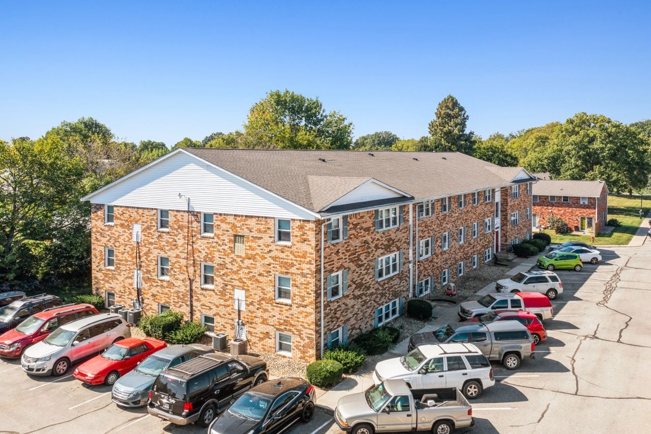 Apartment building with brick exterior, parked cars in front, and trees in background.