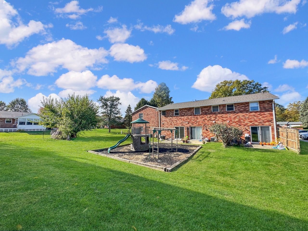 Backyard with brick house, playground, and green grass under a blue sky with fluffy clouds.