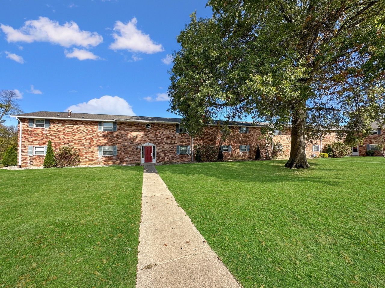Brick apartment building with green lawn, walkway, and a tree under a blue sky.