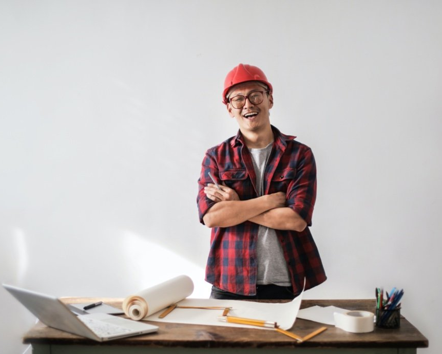 A man wearing a hard hat is standing in front of a desk with his arms crossed.