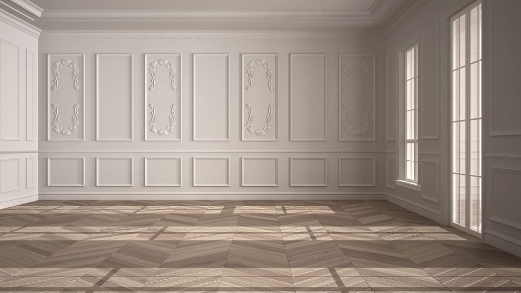 Empty, light-filled room with white paneled walls, ornate carvings, tall windows, and wood parquet flooring.