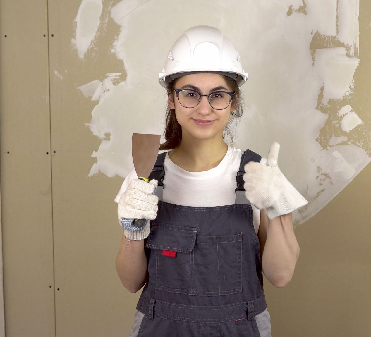 Woman in a hard hat and overalls with a putty knife giving a thumbs-up in front of a drywall patch.