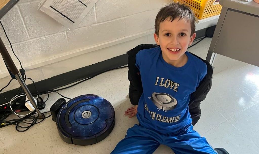 A young boy is sitting on the floor next to a robot vacuum cleaner.