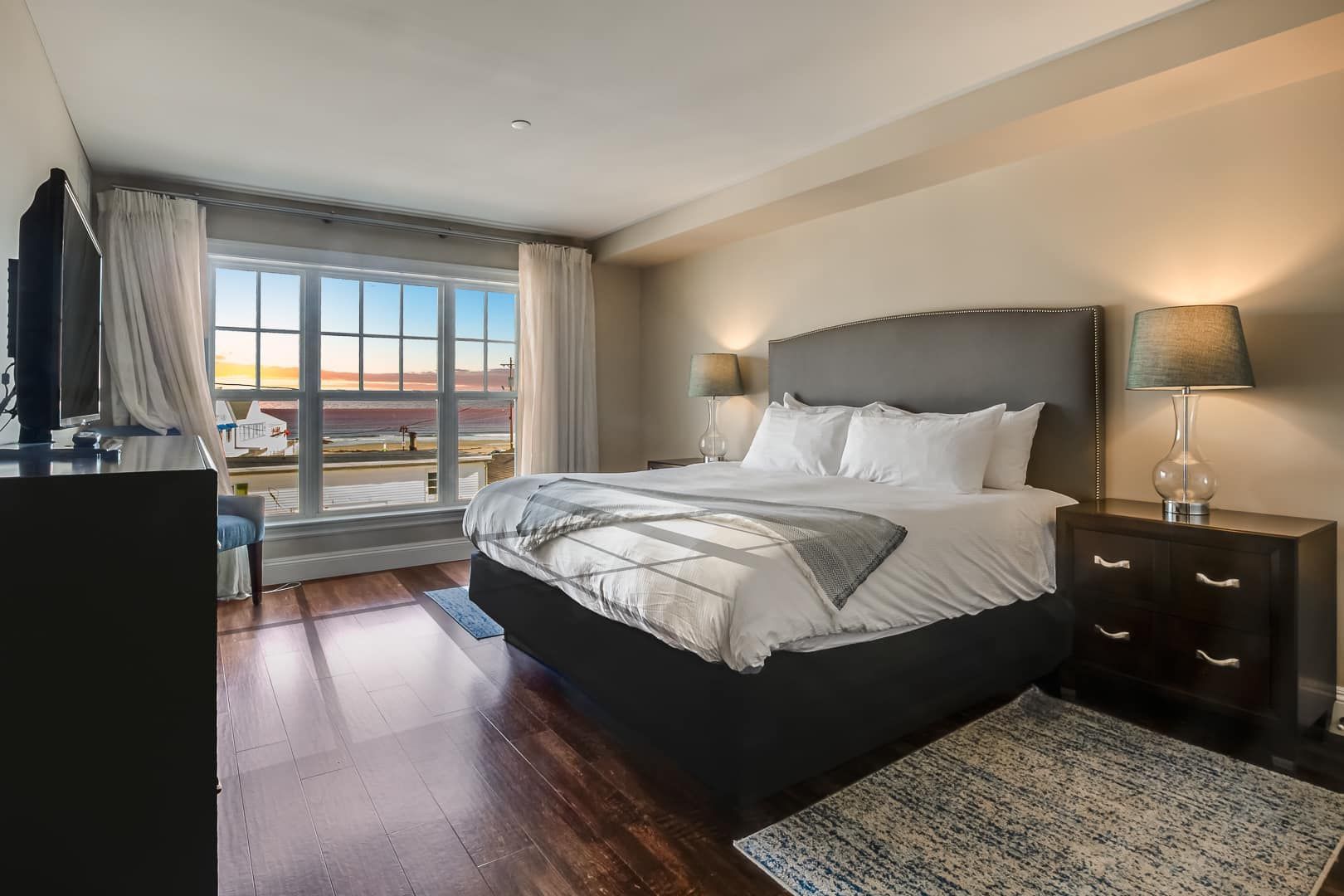 Bedroom with large window overlooking a body of water at sunset, king bed, nightstand with lamp, and rug.