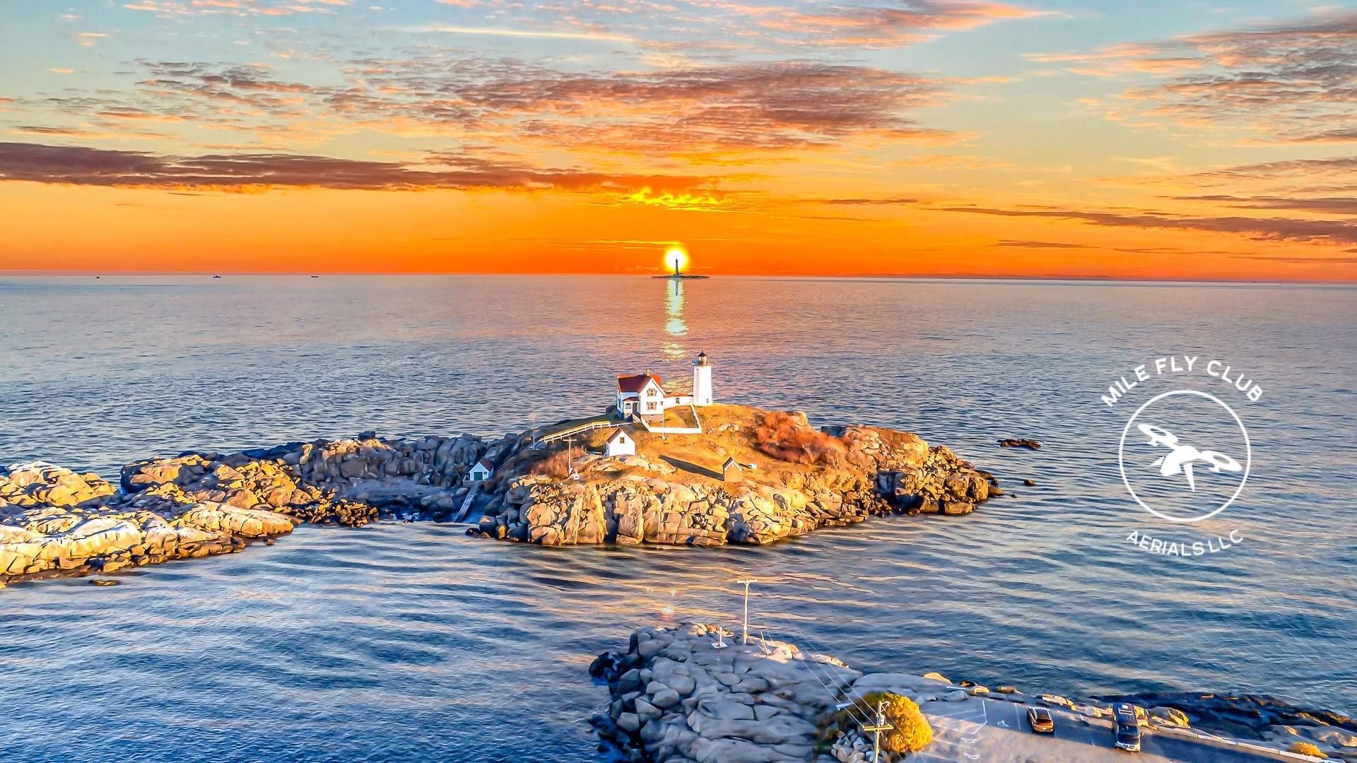 Lighthouse on a rocky island at sunset; orange sky, ocean waves.