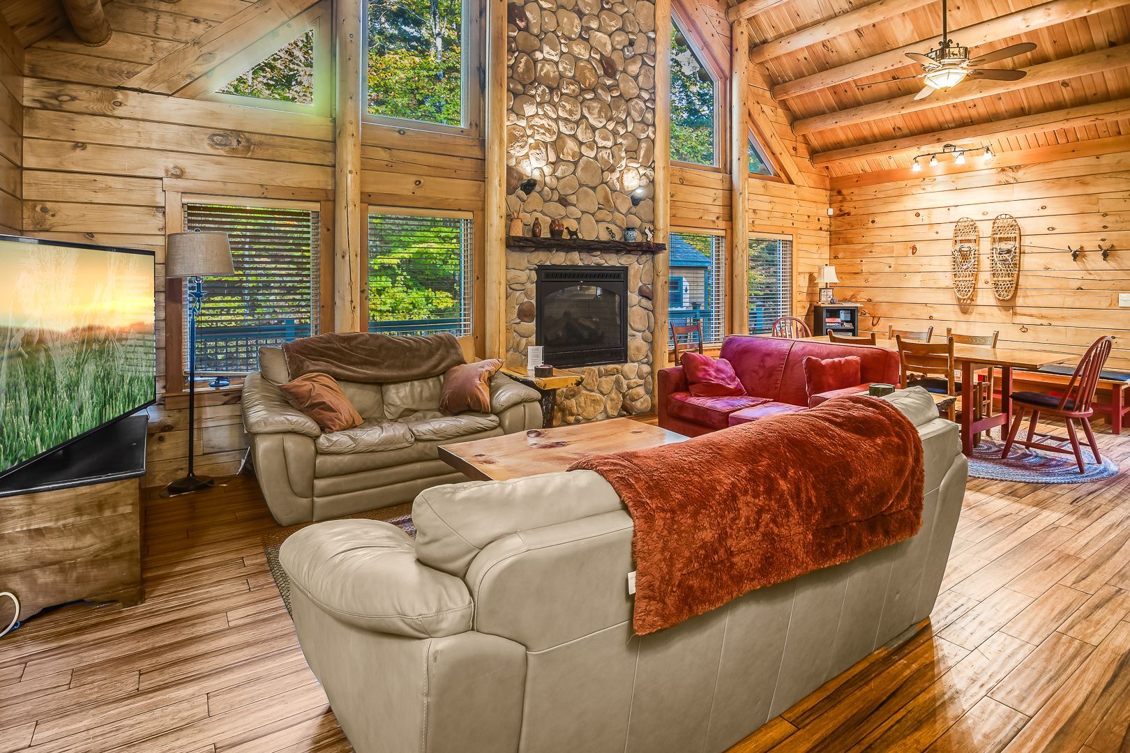 Log cabin living room with stone fireplace, sofas, large windows, and wooden floors.