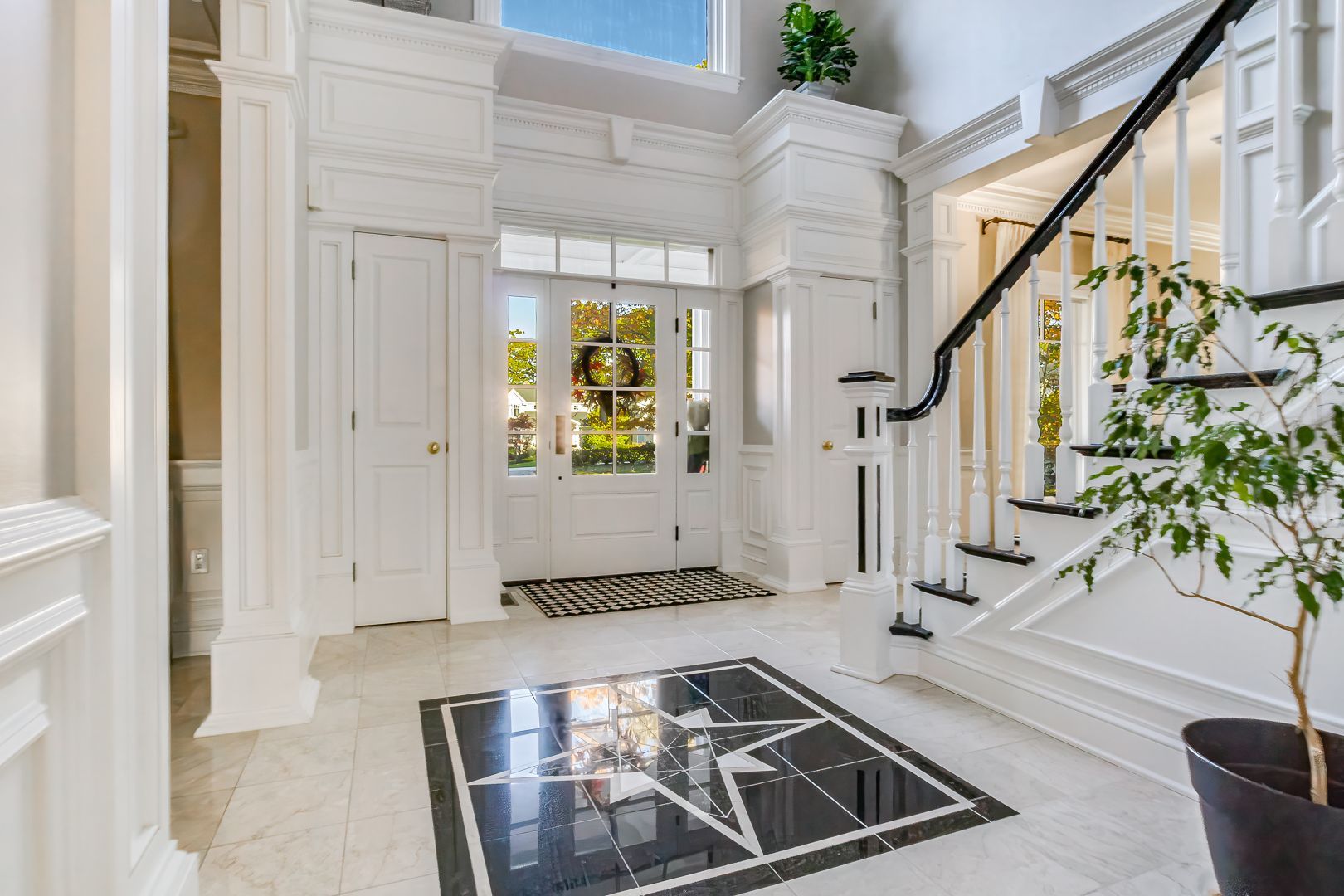 White foyer with black and white tile floor, stairs, and columns. Door in the back.