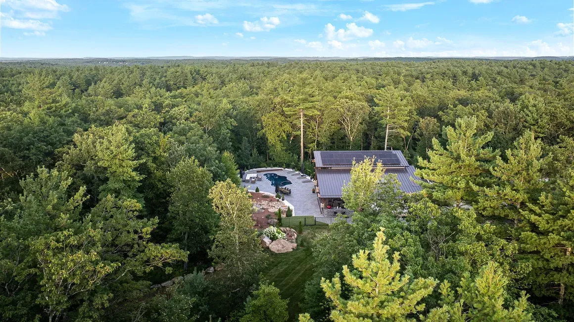 Aerial view of a home surrounded by a dense green forest, with a circular driveway and small yard.