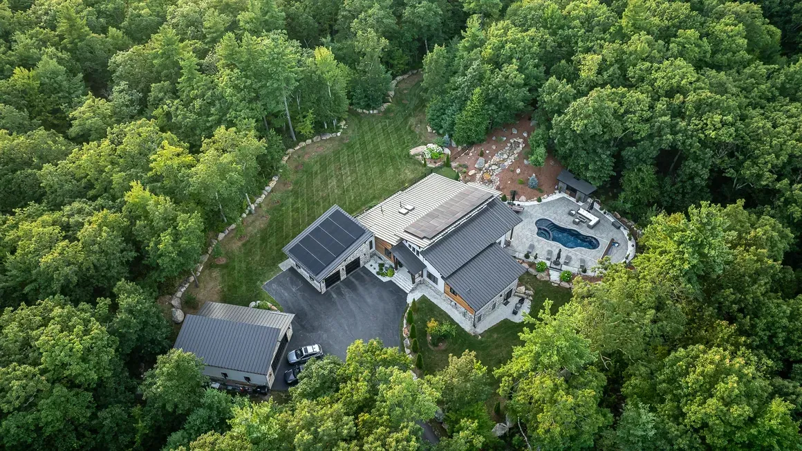 Aerial view of a modern house with a pool and dark roof, surrounded by lush green trees.